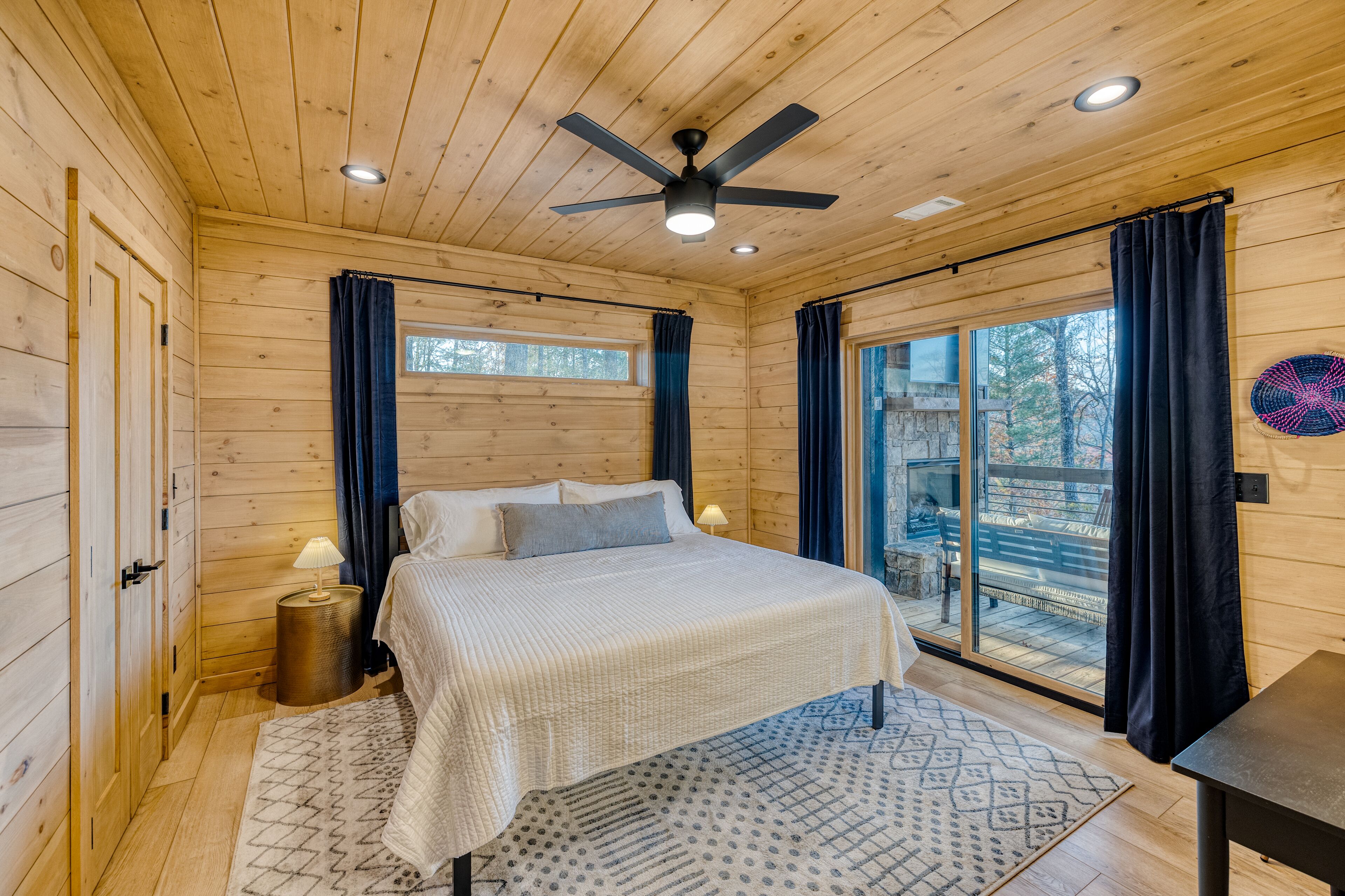A wooden cabin bedroom with a bed, two bedside lamps, a ceiling fan, and a large window with curtains.