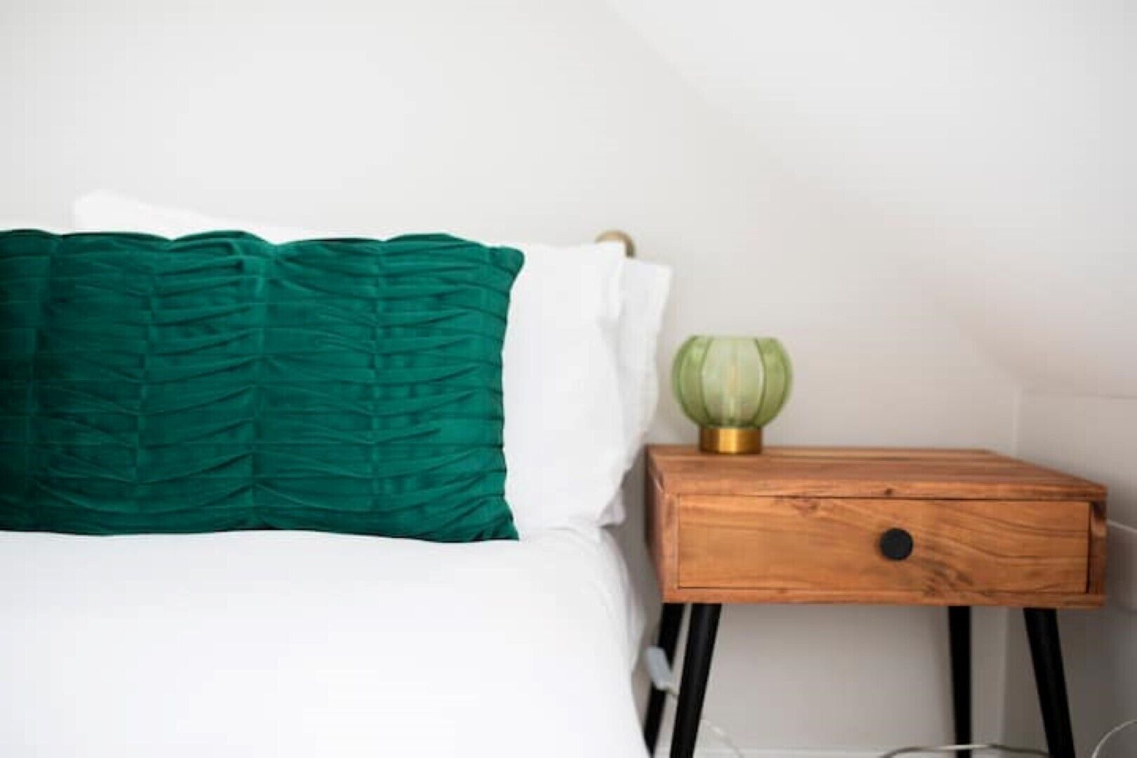 A neatly made bed with a green and white pillow, a wooden nightstand with a green glass lamp, and a drawer.