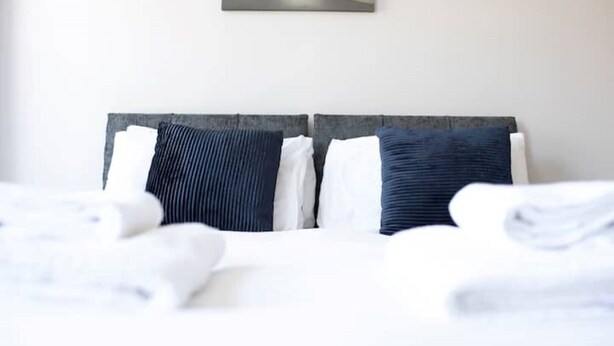 A neatly made bed with white linens and two pillows, one dark and one light, in a room with a plain wall and a painting above the bed.