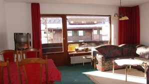 Una sala de estar con un sofá, una mesa y sillas. Hay un televisor sobre un mueble bajo. La habitación tiene una ventana con cortinas y un radiador.