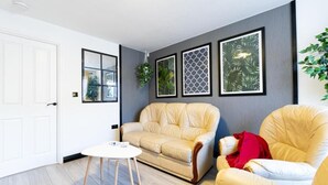 A living room with a beige sofa, a white armchair, and a small round table. There are framed pictures on the wall and a plant hanging above the sofa. - Windy House (Sheffield)