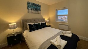 Une chambre à coucher avec un lit, une table de chevet, une lampe, une fenêtre et un tableau au mur.