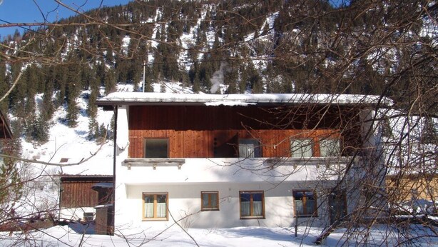 Un chalet à deux étages, avec une façade en bois et des murs blancs, entouré de montagnes et d’arbres couverts de neige.