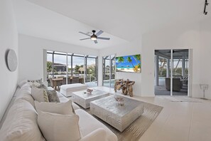 A modern living room with a large sofa, two coffee tables, and a view of a tropical beach through large windows.