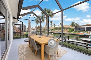 Un espacio de comedor al aire libre cubierto con una mesa y sillas, con vista a un puerto deportivo con barcos y palmeras.