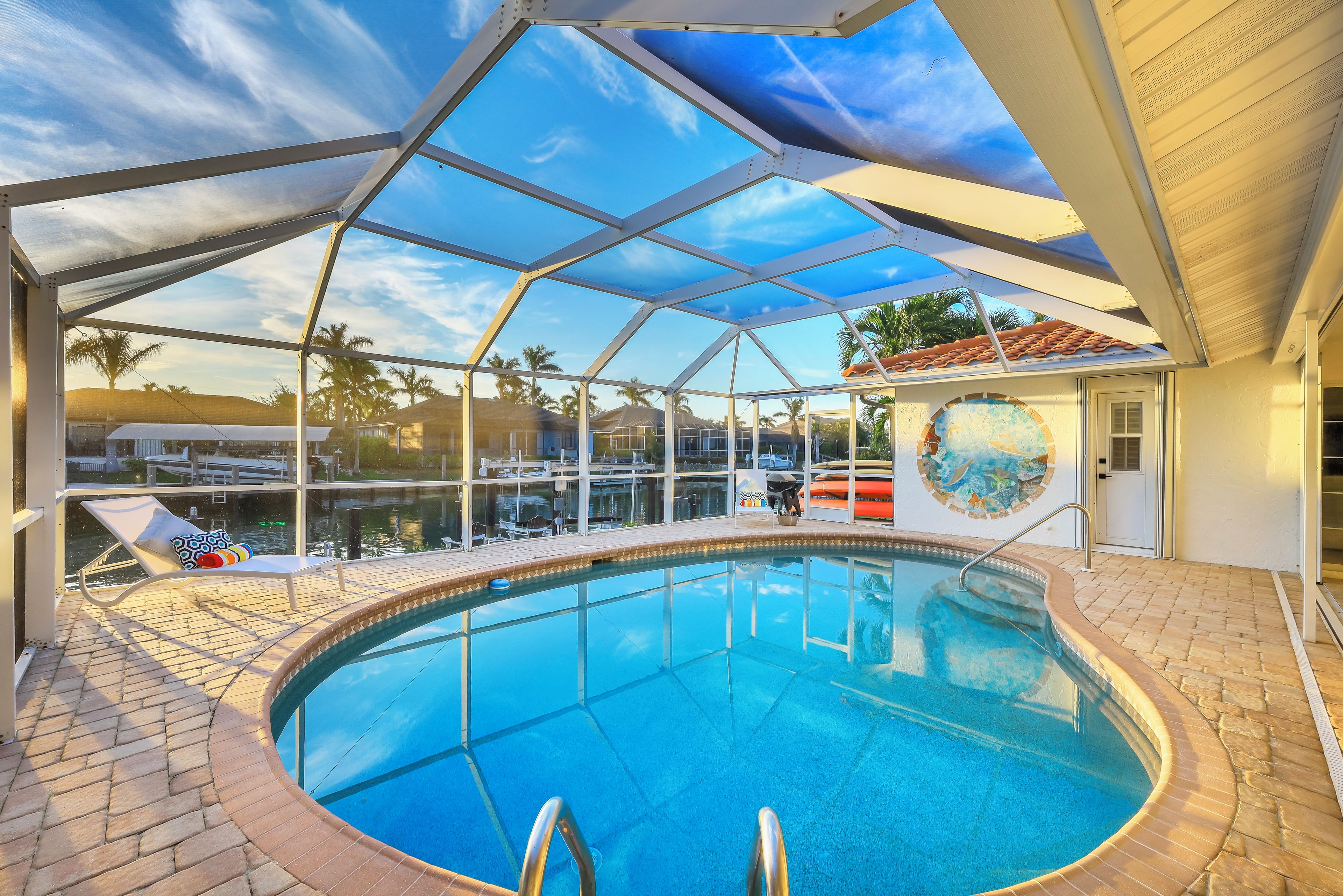 Une piscine entourée de parois en verre, avec des chaises longues et une vue sur les palmiers et les maisons.