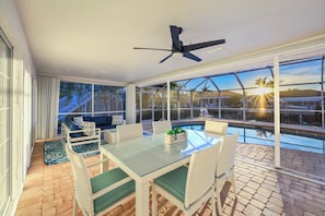 Un patio techado con una mesa de vidrio, sillas de mimbre, un ventilador de techo y vistas a una piscina y al atardecer.