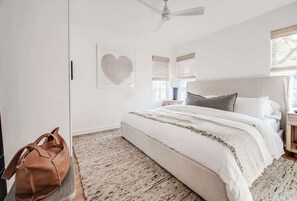 A bedroom with a bed, a ceiling fan, a heart-shaped artwork, and a brown bag on a stand.