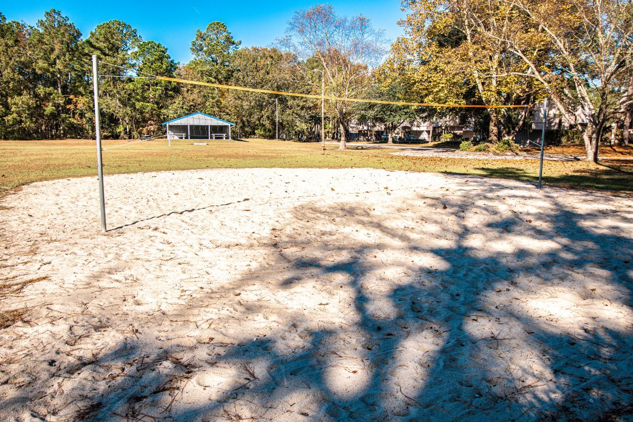 Un terrain de beach-volley sablonneux avec un filet, entouré d’arbres et avec un bâtiment en arrière-plan.