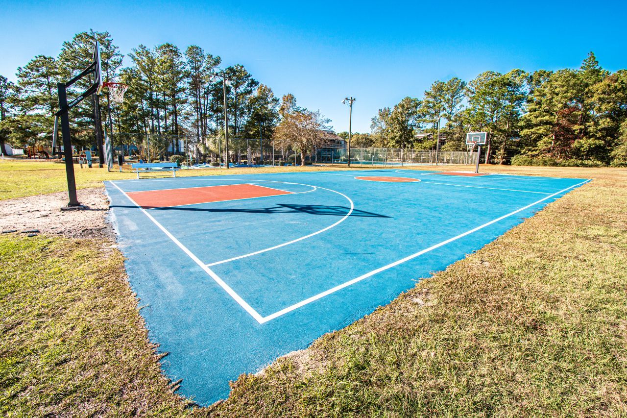 An outdoor basketball court with a blue playing surface, surrounded by grass and trees.
