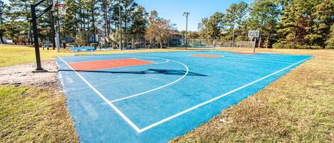 An outdoor basketball court with a blue playing surface, surrounded by grass and trees.