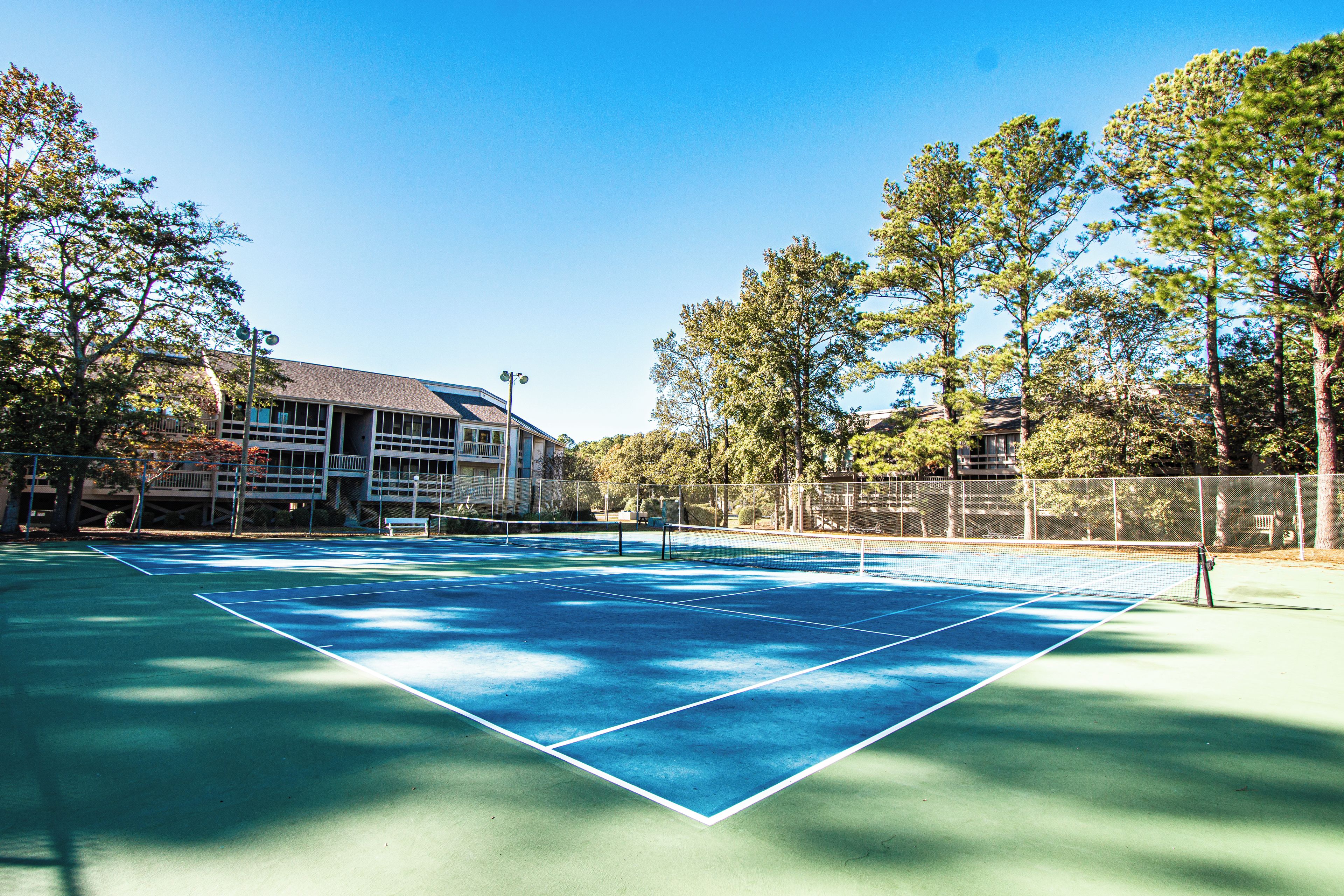 An outdoor tennis court with a net, surrounded by trees and a building in the background.