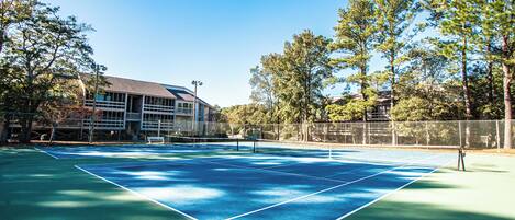 An outdoor tennis court with a net, surrounded by trees and a building in the background.