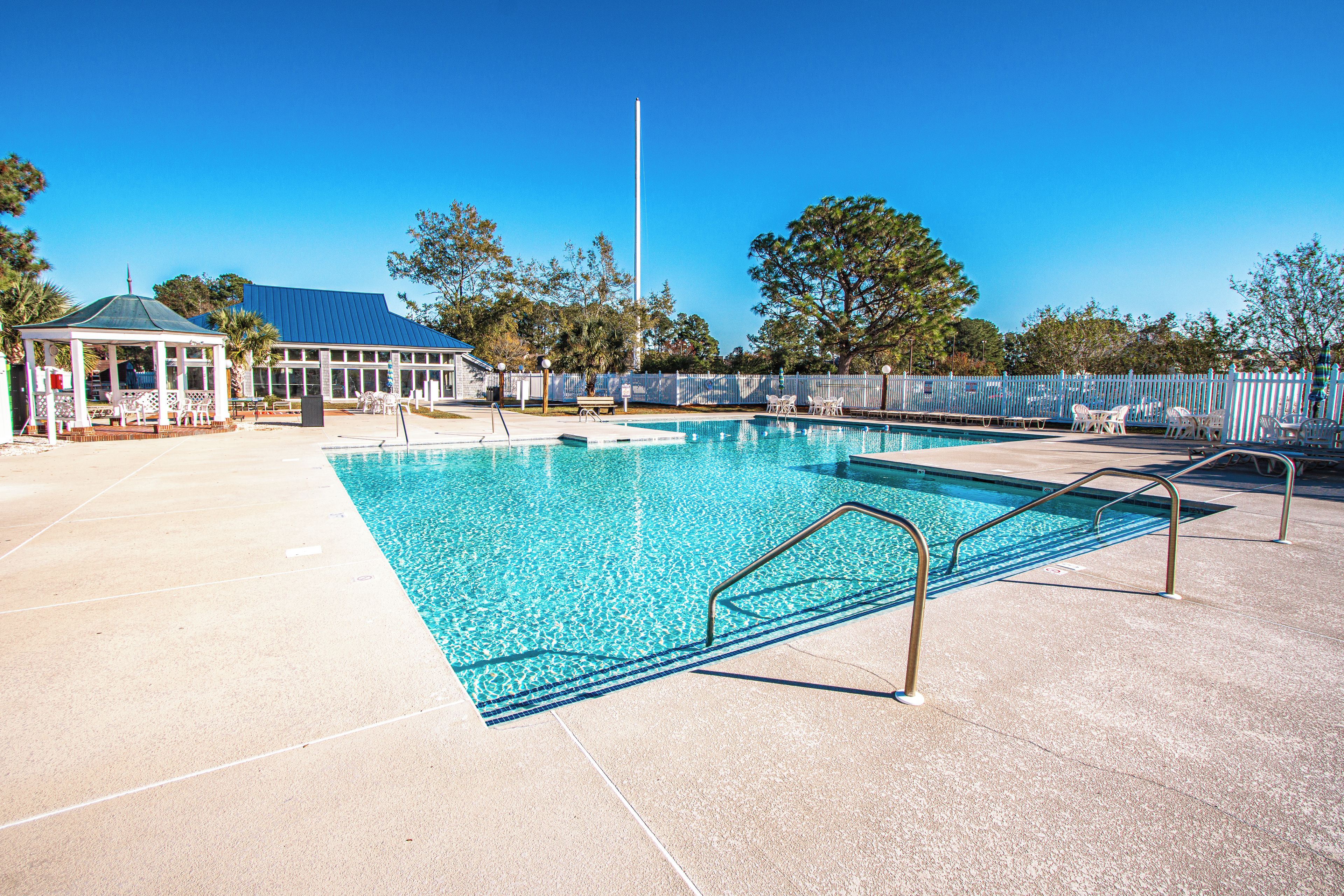 A clear swimming pool with a white fence, lounge chairs, and a gazebo.
