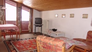 Ein gemütliches Wohnzimmer mit Holzbalkendecke, einem Esstisch mit Stühlen, einer Couch, einem Fernseher auf einem Schrank und einer kleinen weißen Kommode.