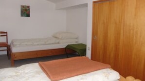 Ein Einzelbett mit Holzrahmen, einem grünen Kissen und einer braunen Decke. Eine Holzliege befindet sich links vom Bett und ein Holzschrank rechts davon.