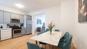 A modern kitchen with white cabinets, stainless steel appliances, and a marble countertop. There is a dining area with teal chairs and a vase of flowers.