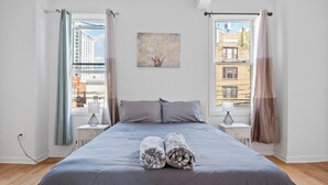 A bedroom with a large bed, two bedside tables, a window with curtains, and a city view.