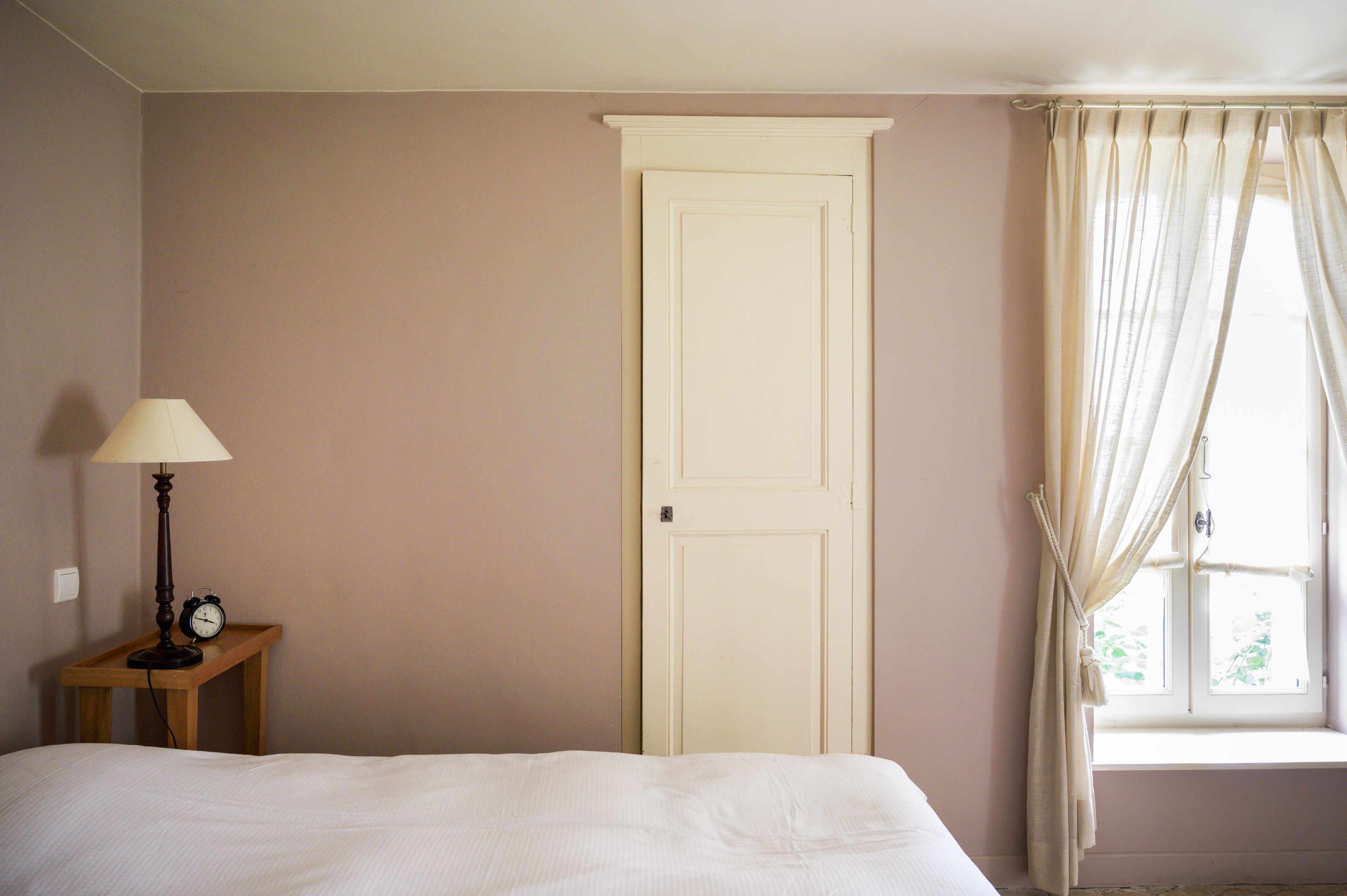 A bedroom with a bed, a nightstand with a lamp and an alarm clock, a door, and a window with curtains.