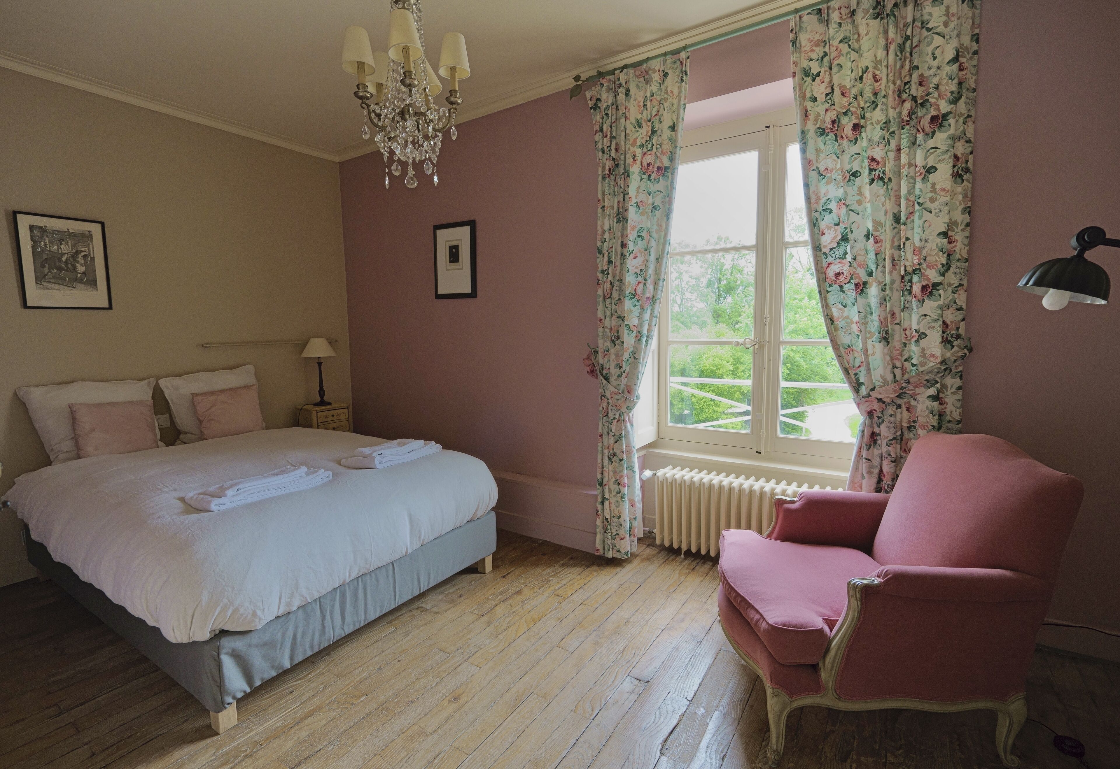 A bedroom with a bed, a chair, a window with floral curtains, and a chandelier.