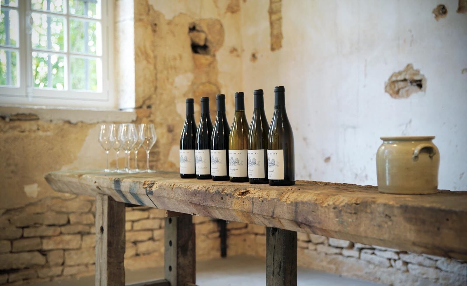A rustic wooden table with wine bottles and glasses in a room with stone walls and a window.
