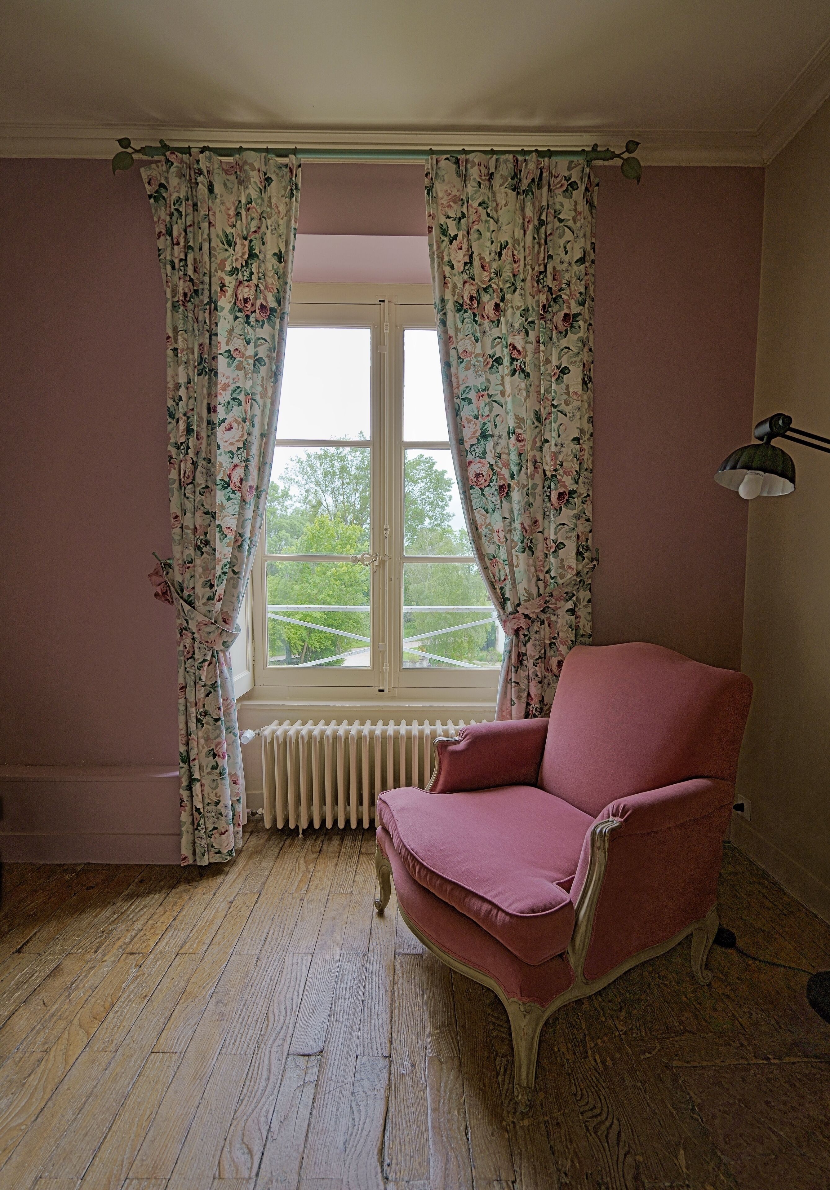 A room with a pink chair, floral curtains, a radiator, and a window with a view of greenery.