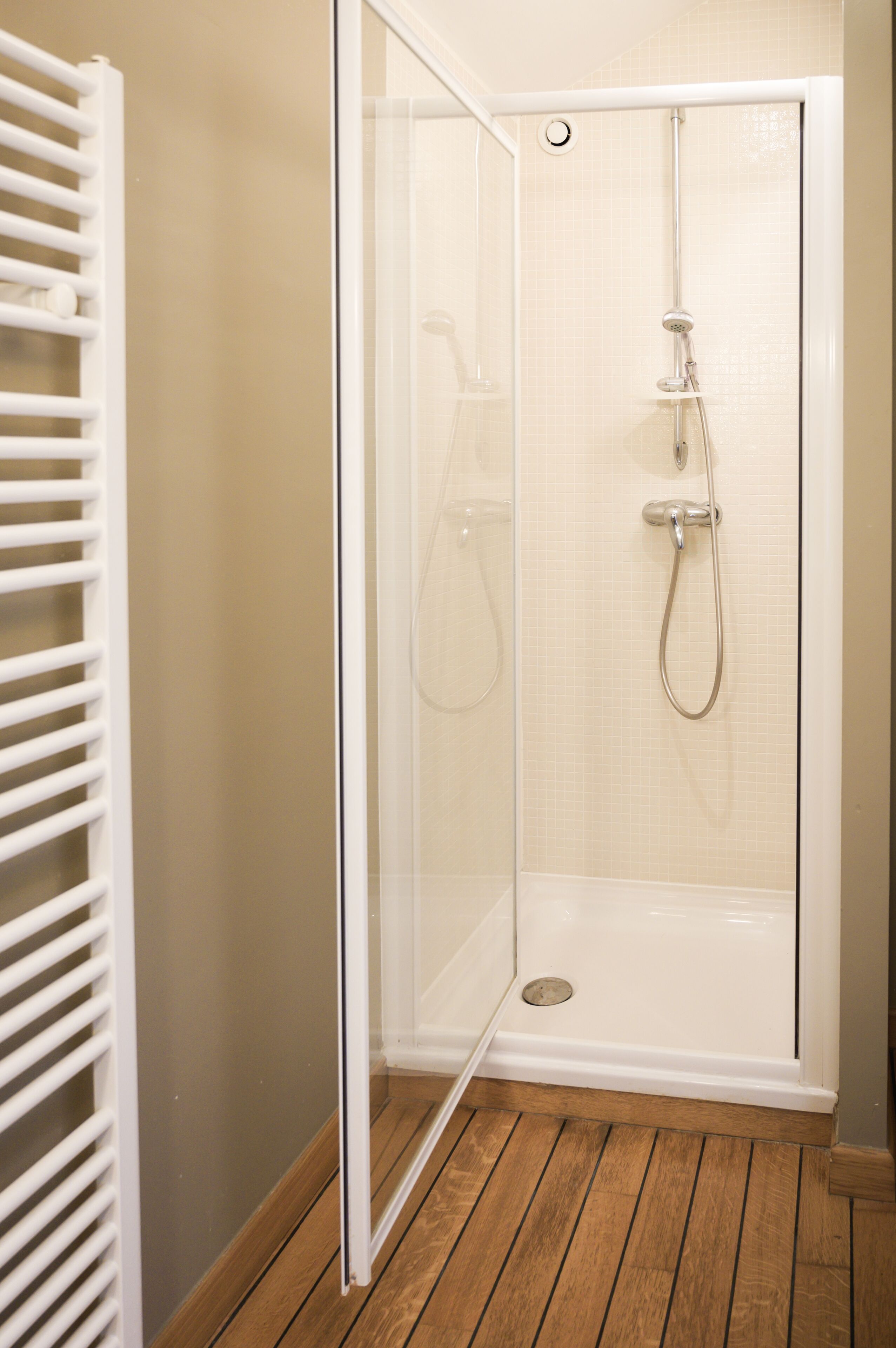 Ducha a pie con puerta de vidrio, suelo de madera y un toallero blanco.