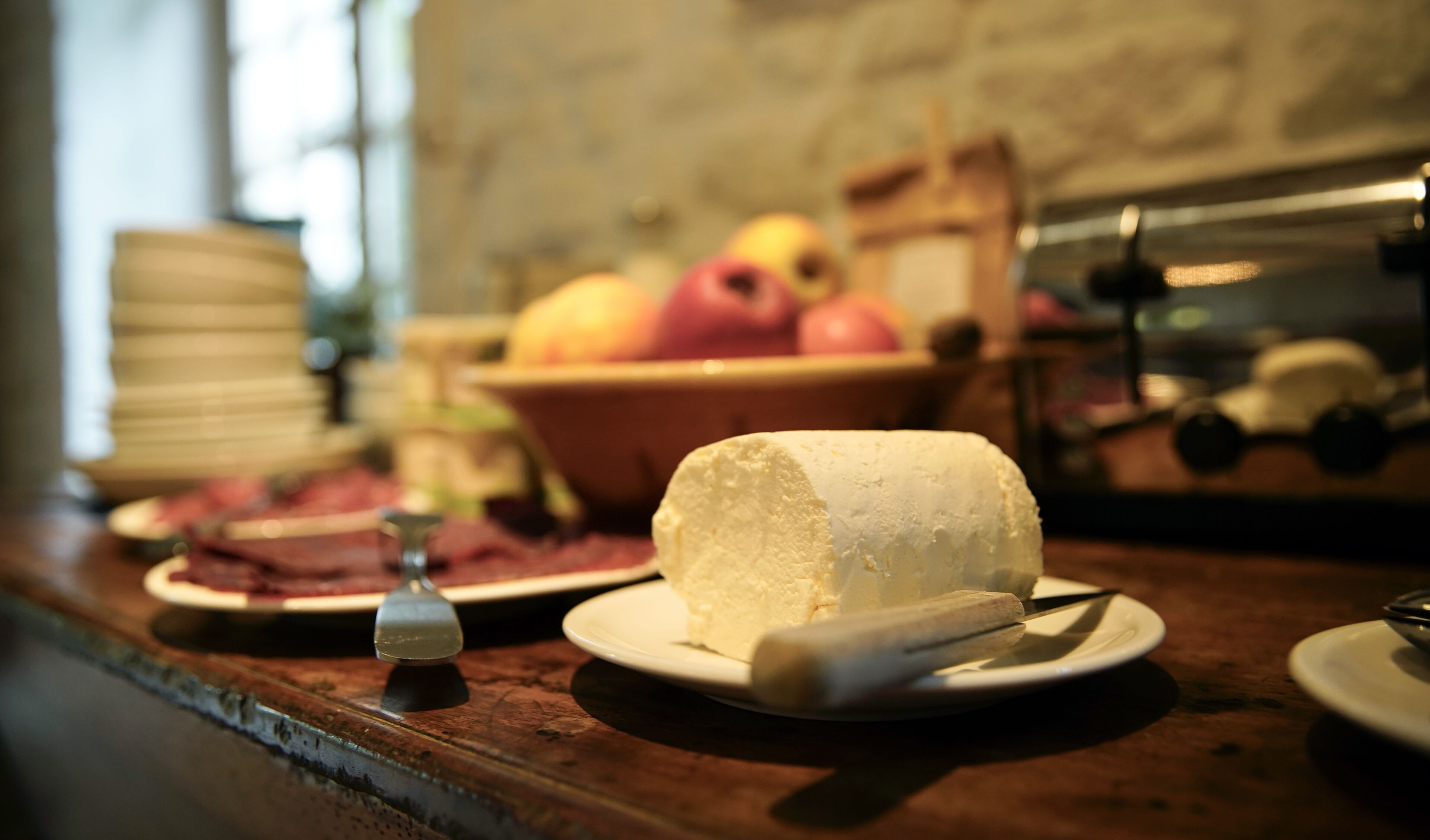 Une portion de fromage présentée sur une assiette, avec un couteau à proximité, un bol de fruits et une pile d’assiettes en arrière-plan.