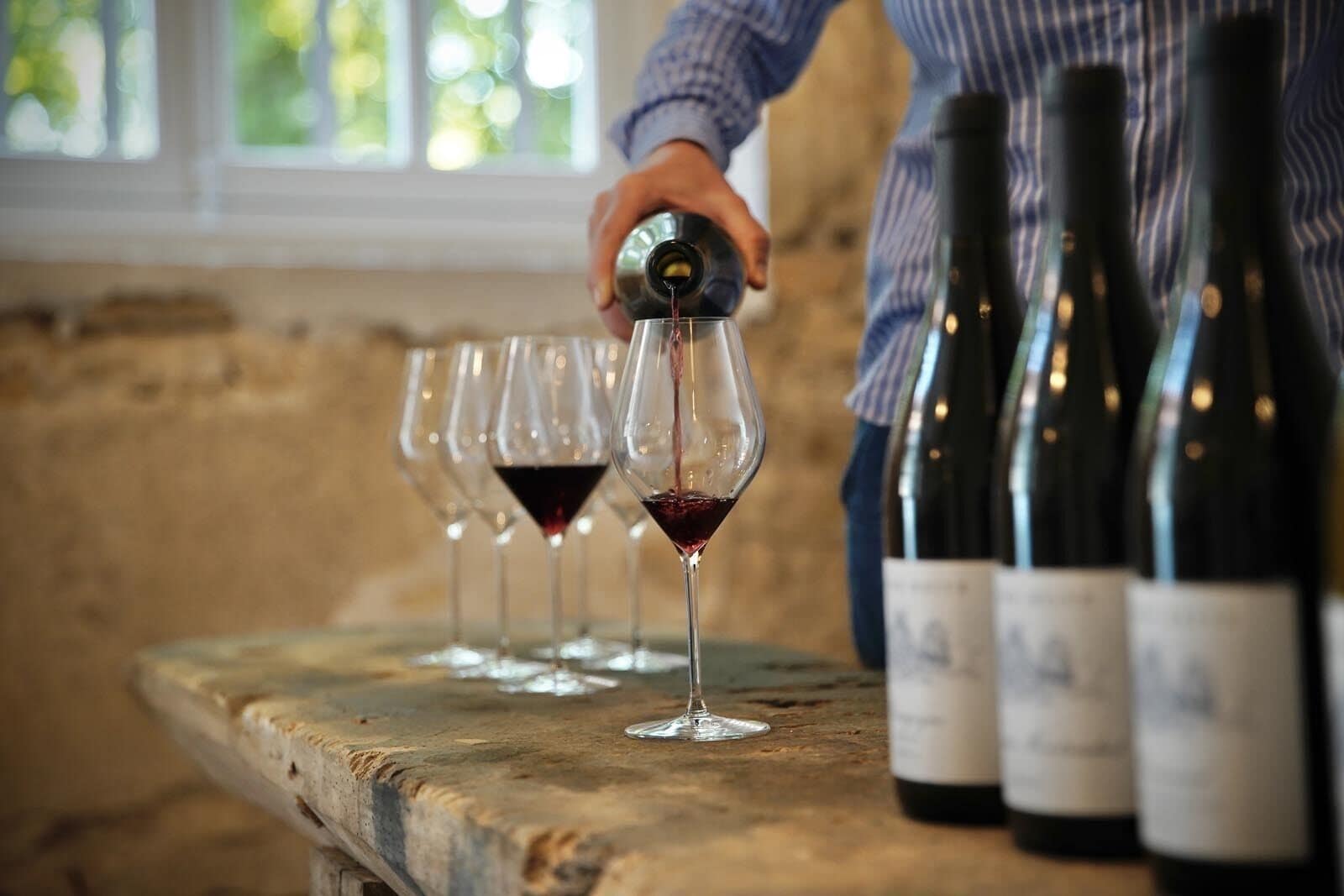 Eine Person gießt Wein in Gläser auf einem rustikalen Tisch, im Hintergrund stehen Flaschen.
