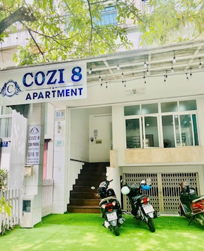 Ein Mehrfamilienhaus namens COZI 8 mit Vermietungshinweis. Vor dem Eingang führen Treppen hinauf, und mehrere Motorräder sind geparkt.
