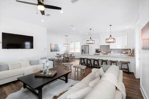 A spacious living room with a sofa, coffee table, and a TV mounted on the wall. There is a dining area with a table and chairs, and a kitchen with bar stools.