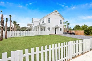 A large white two-story house with a front porch, surrounded by a white picket fence, palm trees, and a clear blue sky. - Sundance: Sleeps 18! 1 Block to Beach! Pool & spa! (Destin)