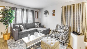 Un salon avec un canapé d’angle gris, un fauteuil à motifs, une table basse, une plante en pot, une horloge murale et un rideau à rayures.