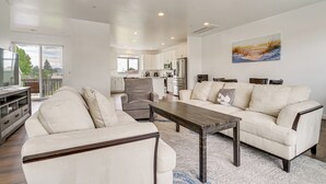 Ein modernes Wohnzimmer mit Sofa, Ohrensessel und Couchtisch, im Hintergrund eine Küche und ein Gemälde an der Wand.
