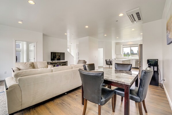 A modern living room with a large sofa, dining table, and chairs, a kitchen in the background, and a large window.