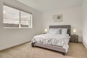 A bedroom with a large window, a bed with a grey headboard, two bedside tables with lamps, and a wall-mounted artwork.