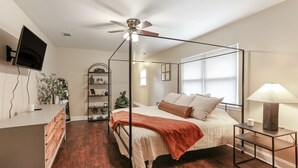 Un dormitorio con cama con dosel, un escritorio con silla, una lámpara de pie y un televisor montado en la pared.