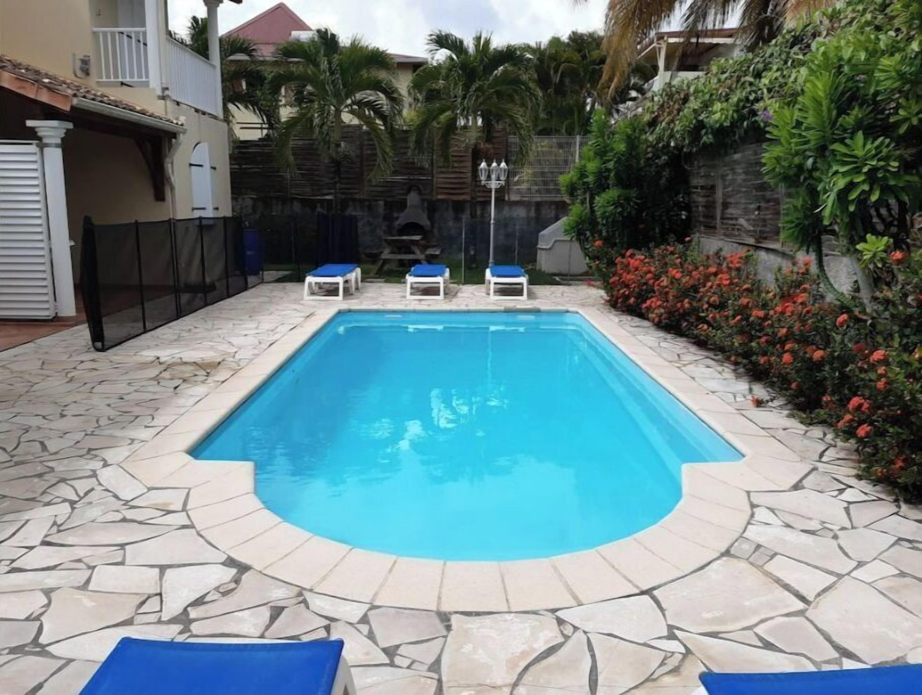 A rectangular swimming pool with a stone border, surrounded by a stone patio. There are three lounge chairs with blue cushions.
