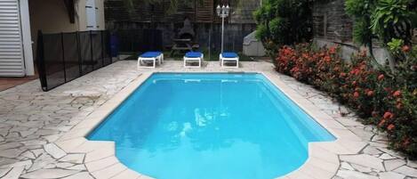 A rectangular swimming pool with a stone border, surrounded by a stone patio. There are three lounge chairs with blue cushions.