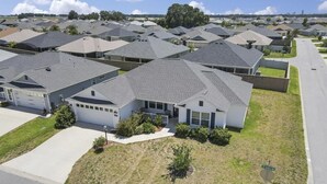 Zona residencial con casas de techos grises y fachadas blancas, dispuestas en un patrón de cuadrícula, con céspedes cuidados y cielo despejado.