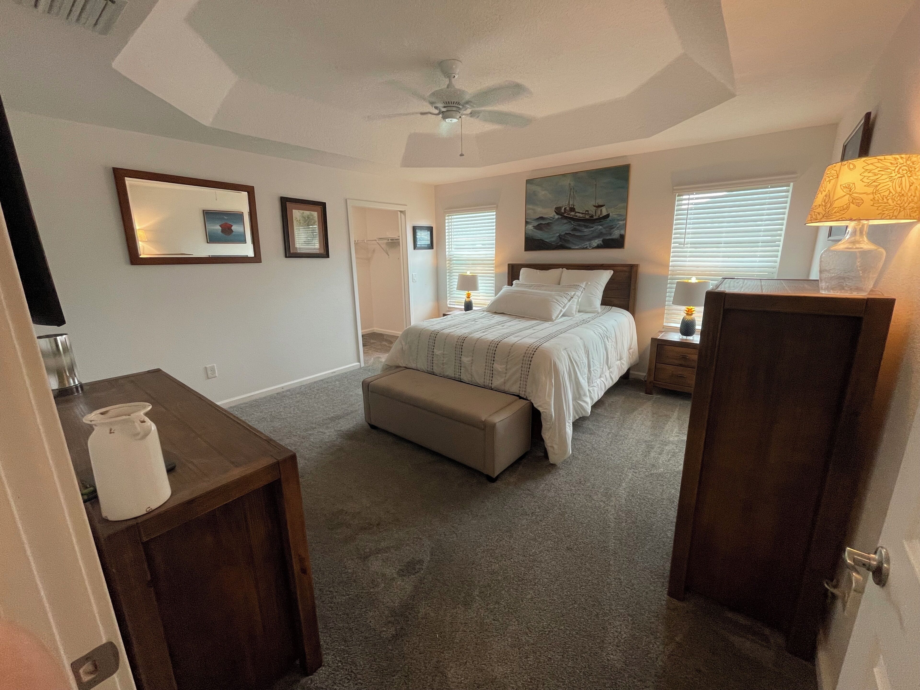 A bedroom with a bed, a desk, a chair, a nightstand, a lamp, a ceiling fan, and a painting on the wall.