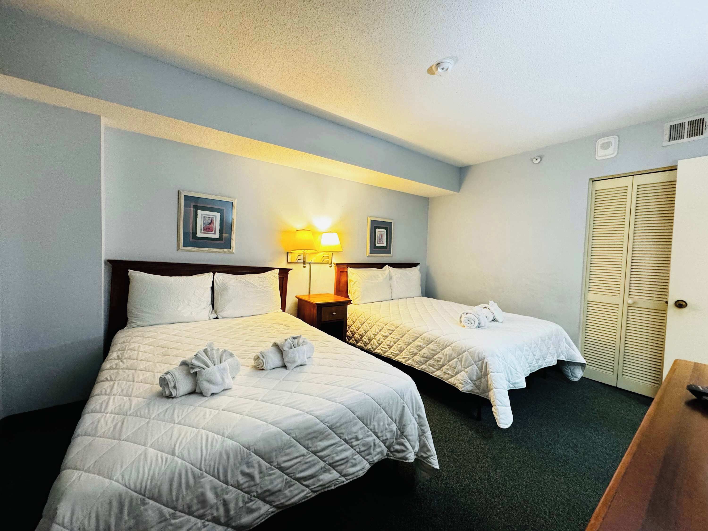 A hotel room with two beds, a nightstand with a lamp, a wooden wardrobe, and a carpeted floor.