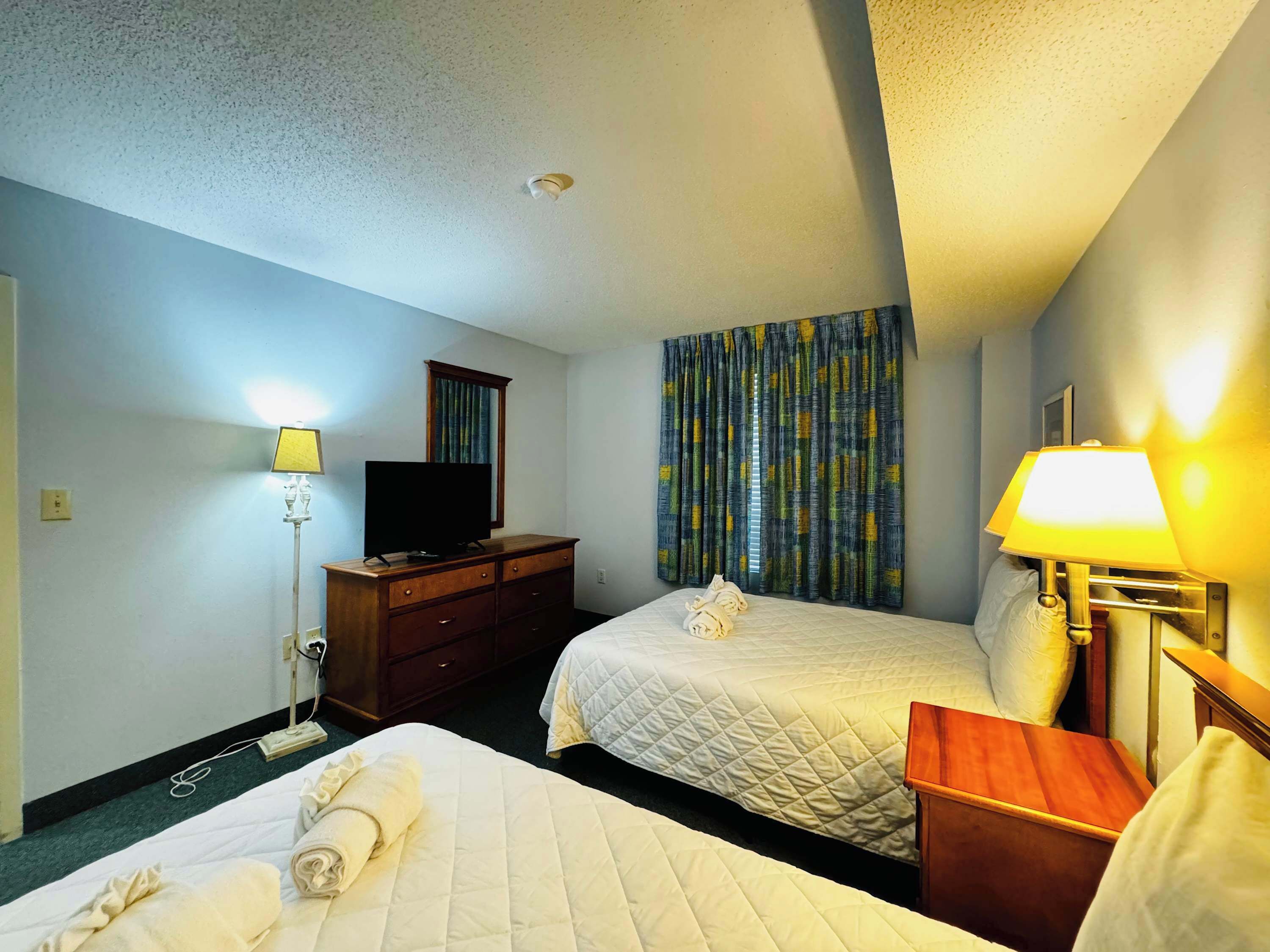 A hotel room with two beds, a television on a dresser, a lamp, and a window with curtains.
