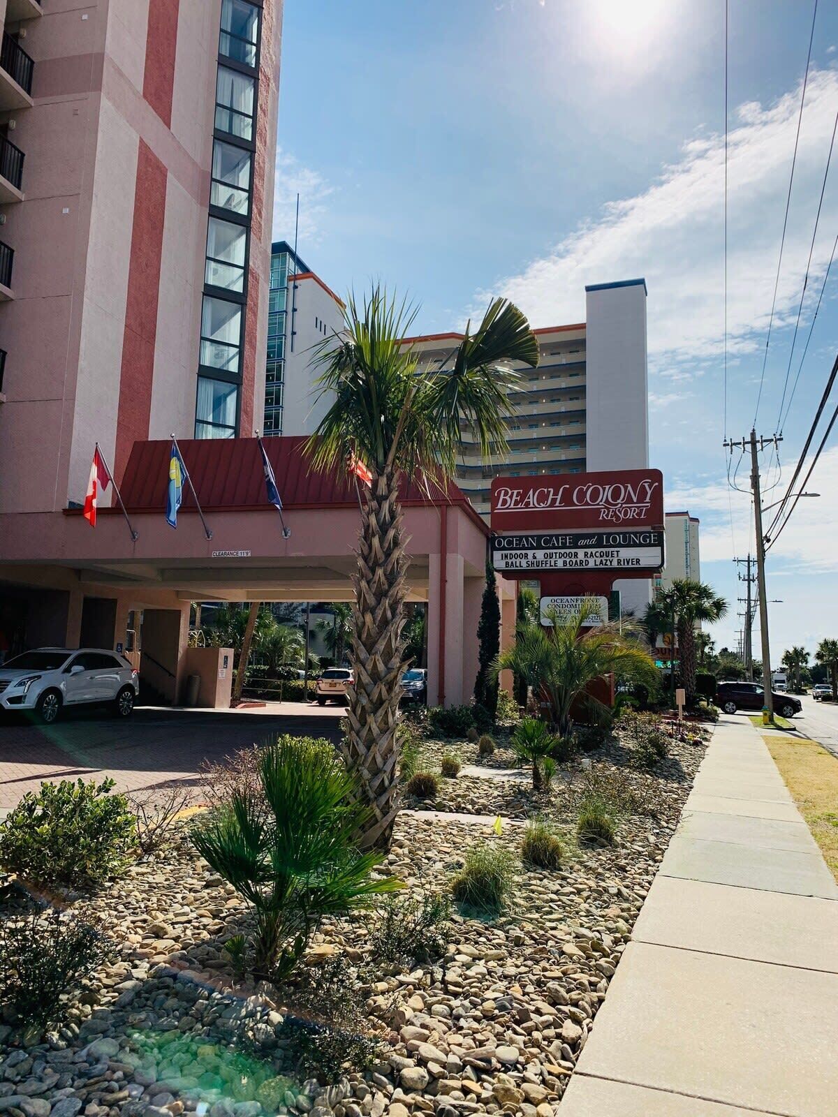 A hotel with a sign for 'Beach Colony Resort' featuring amenities like an ocean cafe, lounge, indoor and outdoor racquet, and board lazy river.
