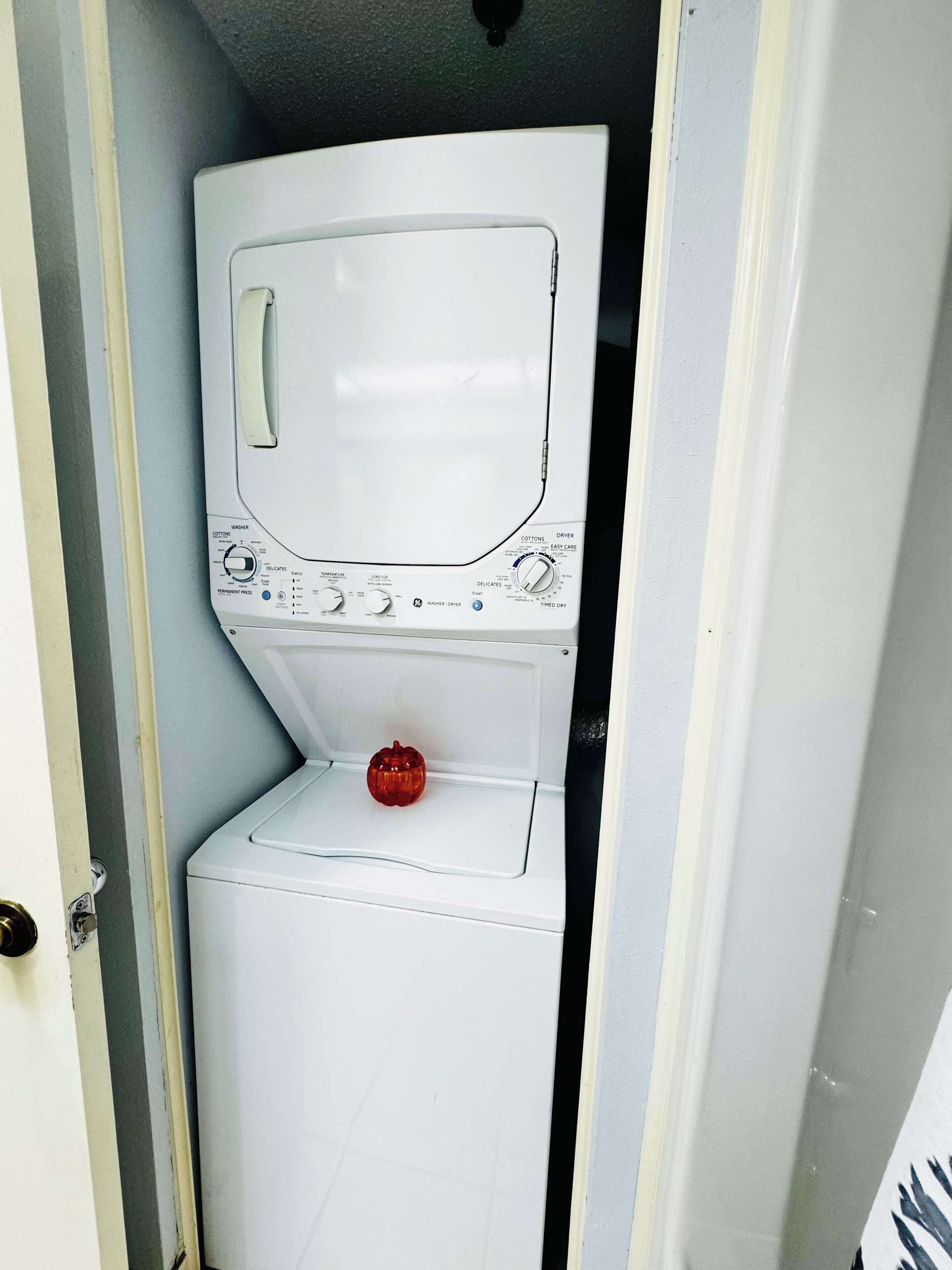 Una lavadora blanca y compacta en un pequeño cuarto de lavado cerrado, con una puerta a un lado.