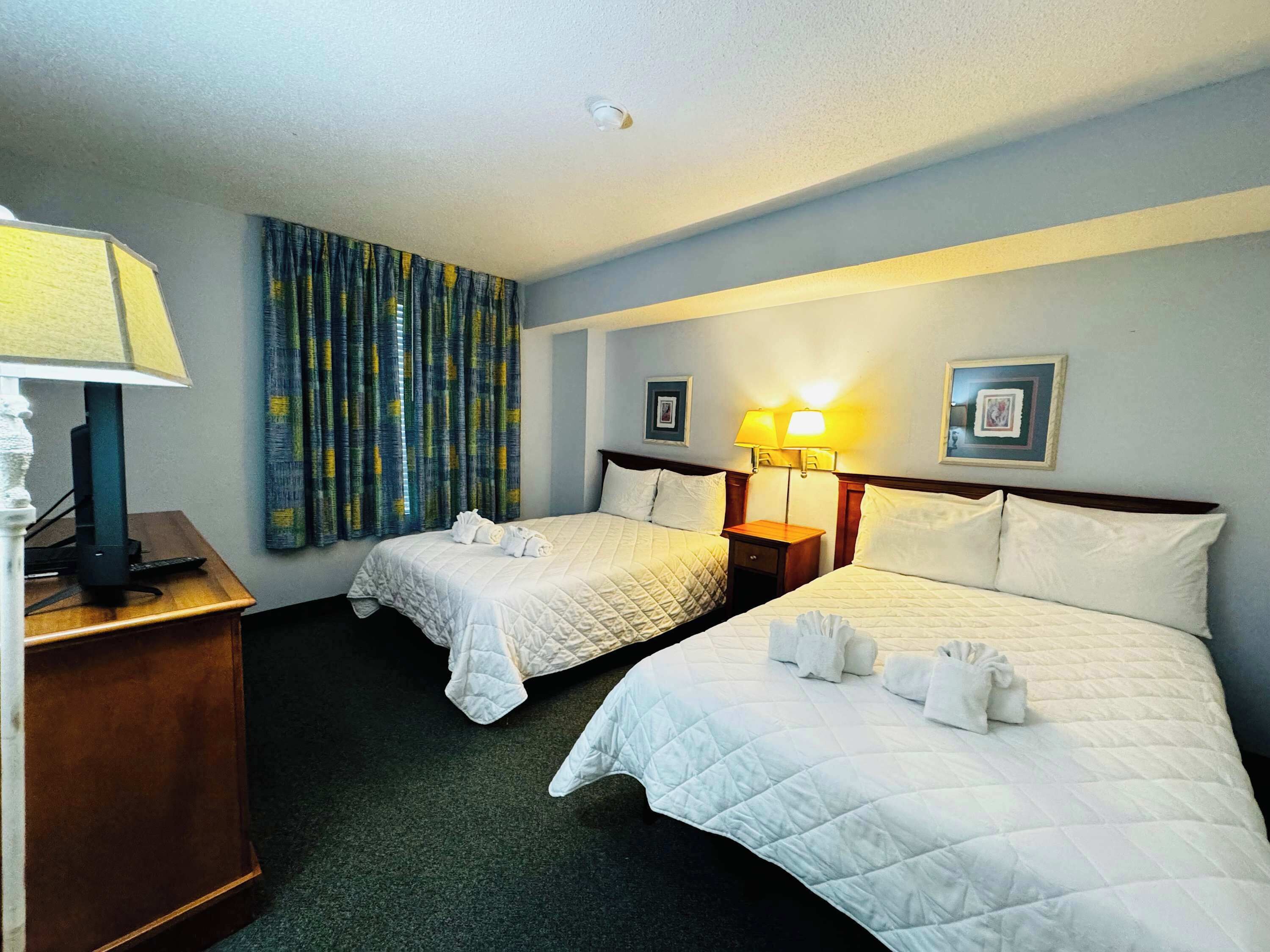 A hotel room with two beds, a desk with a lamp, and a window with curtains.