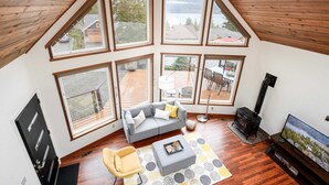 A loft-style room with a sloped ceiling, large windows, a gray sofa, a yellow armchair, a fireplace, and a television.