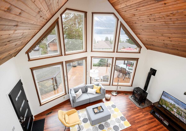 A loft-style room with a sloped ceiling, large windows, a gray sofa, a yellow armchair, a fireplace, and a television.