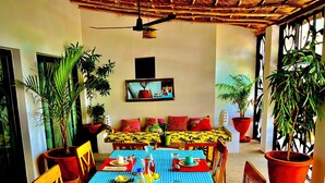 A dining area with a table set for two, surrounded by chairs, potted plants, and a sofa with cushions. - Bine Bassile (Guereo)