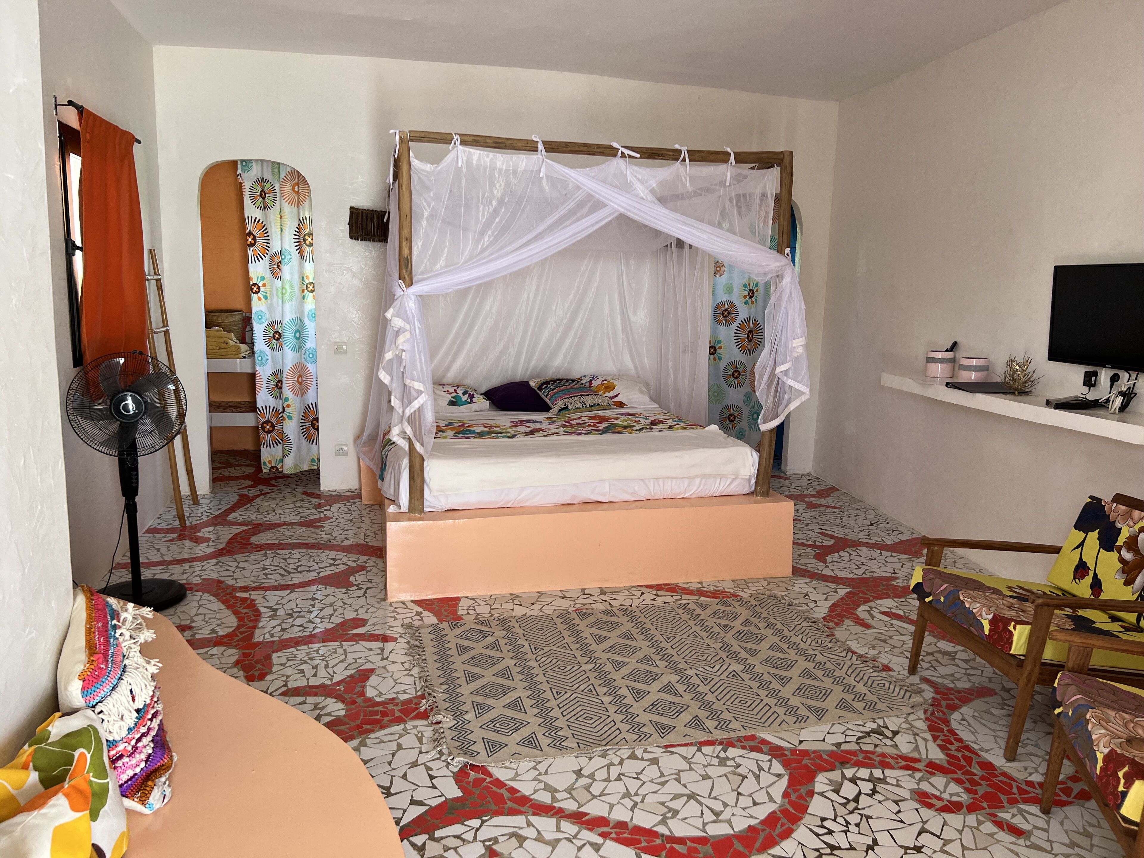 Un dormitorio con una cama bajo un dosel, un televisor en un estante, un ventilador y un suelo estampado.
